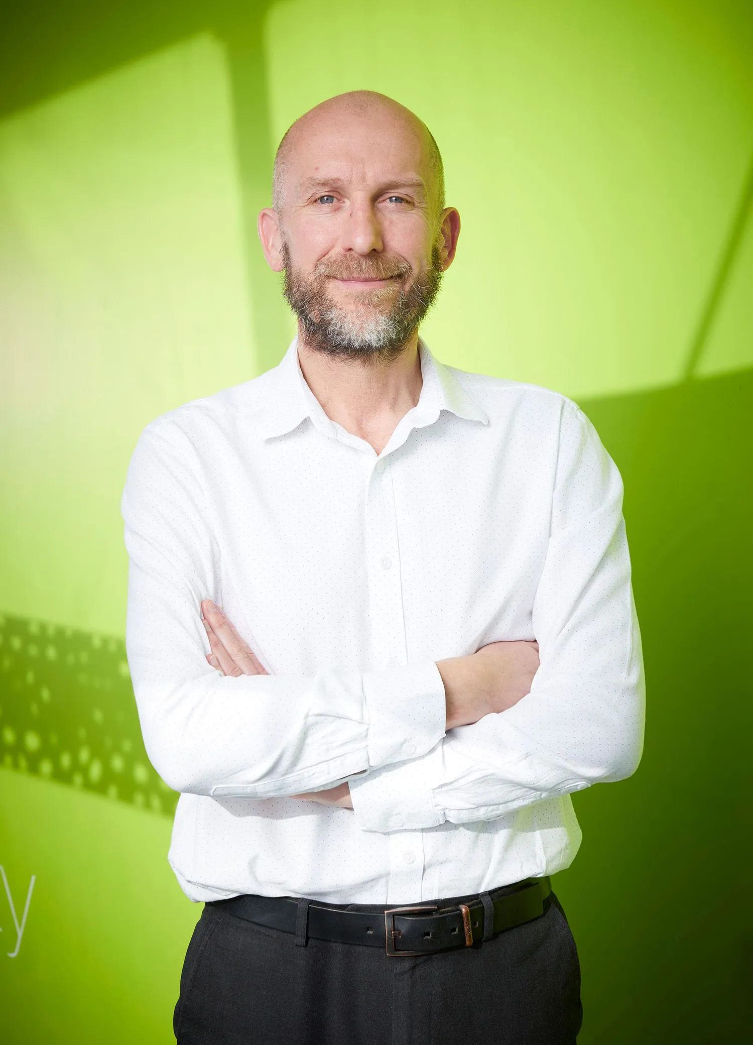 Bald, bearded man in a white shirt with folded arms against a lime-green wall