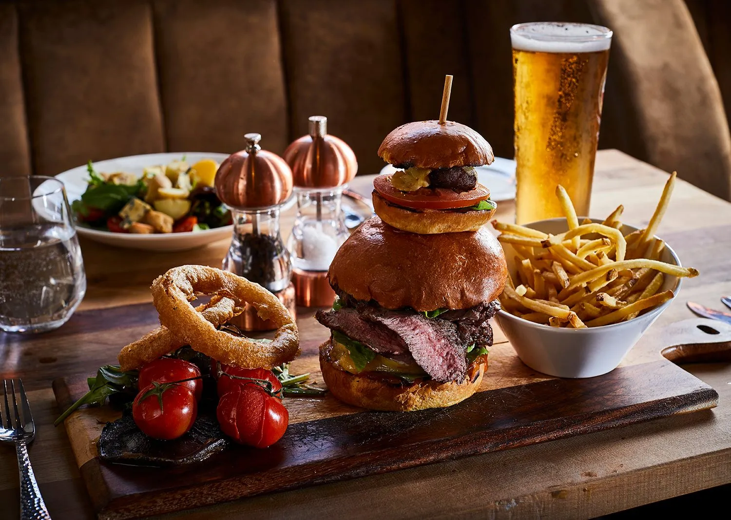 Stacked steak burger with onion rings, fries and a pint of lager on a pub table