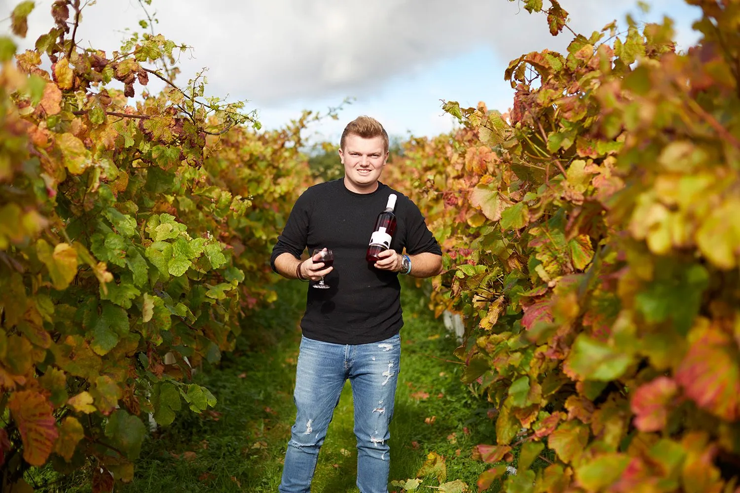 Winemaker holding a bottle and glass between autumn vines