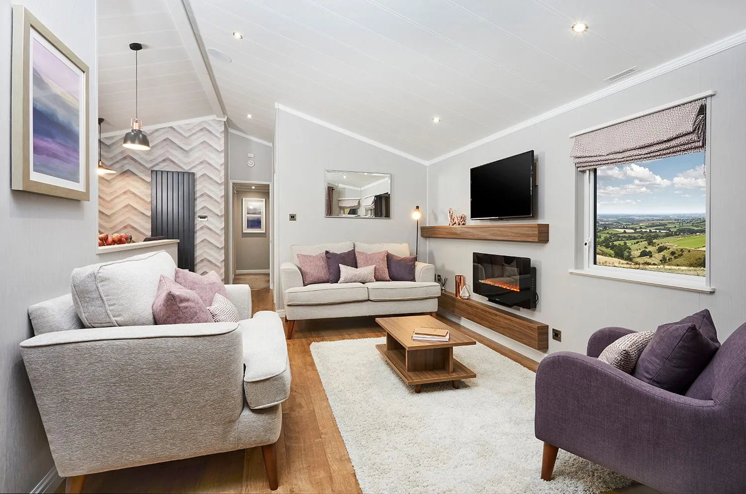 Holiday lodge lounge with grey sofas, a log-effect fire and a window looking over green hills