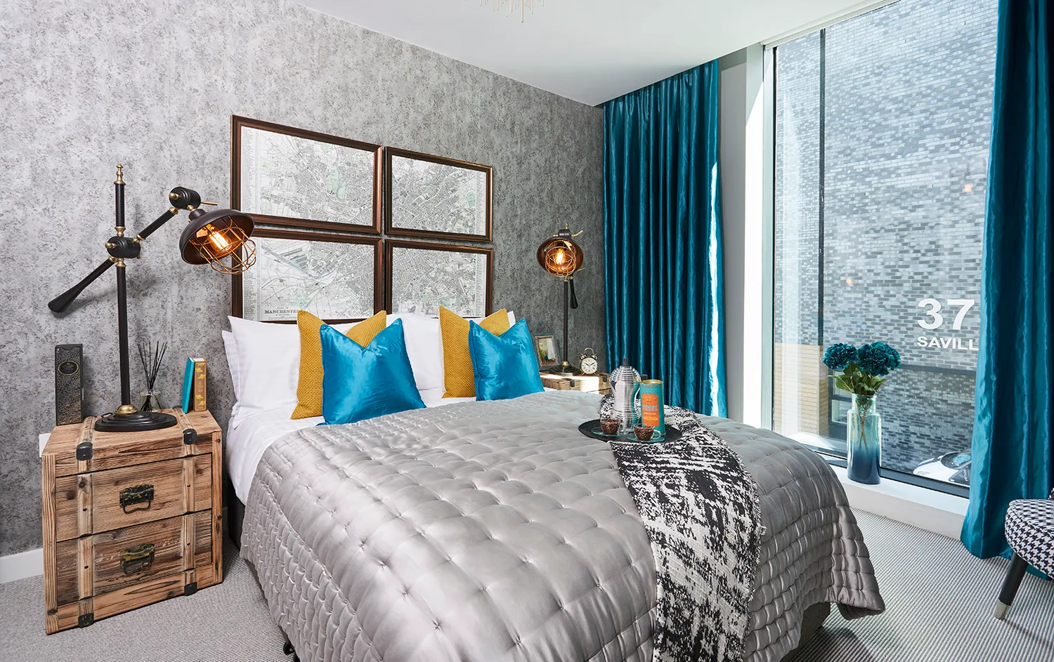 Apartment bedroom with a quilted grey bedspread, teal cushions and full-height windows