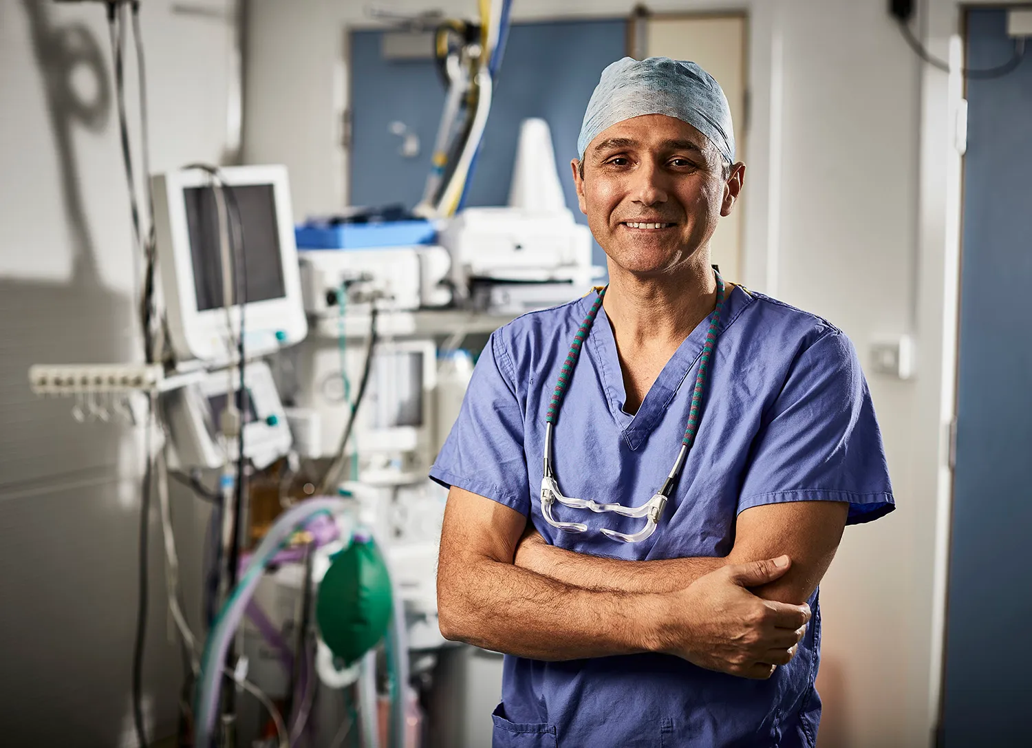 Surgeon in blue scrubs and a theatre cap, arms folded, in an operating theatre